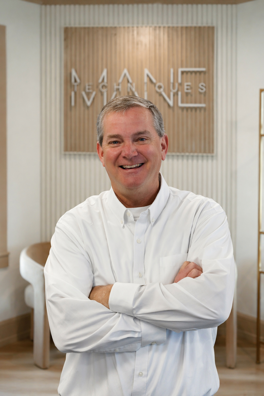 Smiling man in a white button-down shirt stands with arms crossed in a modern reception area with a wood-paneled backdrop behind him.